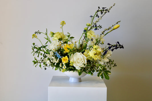 Buttercream and meadow-inspired floral arrangement with soft yellow, white, and blue blooms in a low ceramic vase.