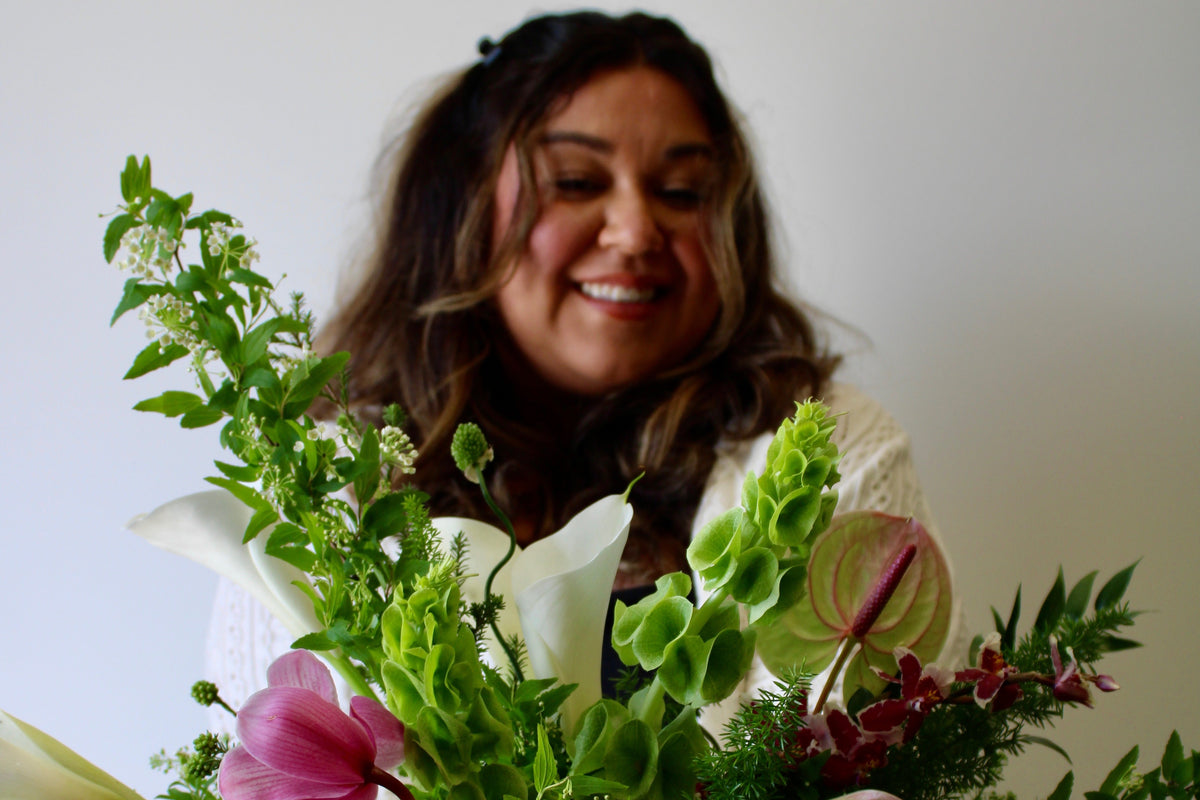 A smiling woman holding a large, vibrant floral bouquet with pink flowers, white lilies, and green foliage against a plain light gray wall.